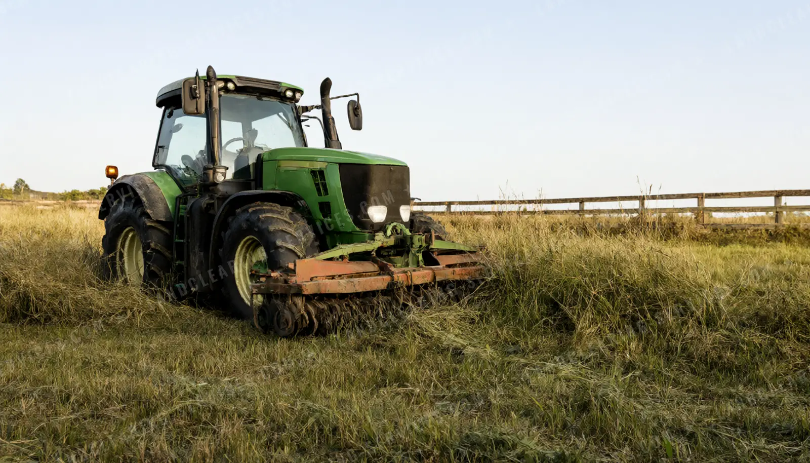 Green tractor bush hogging an overgrown pasture along a wooden fence line, bush hogging