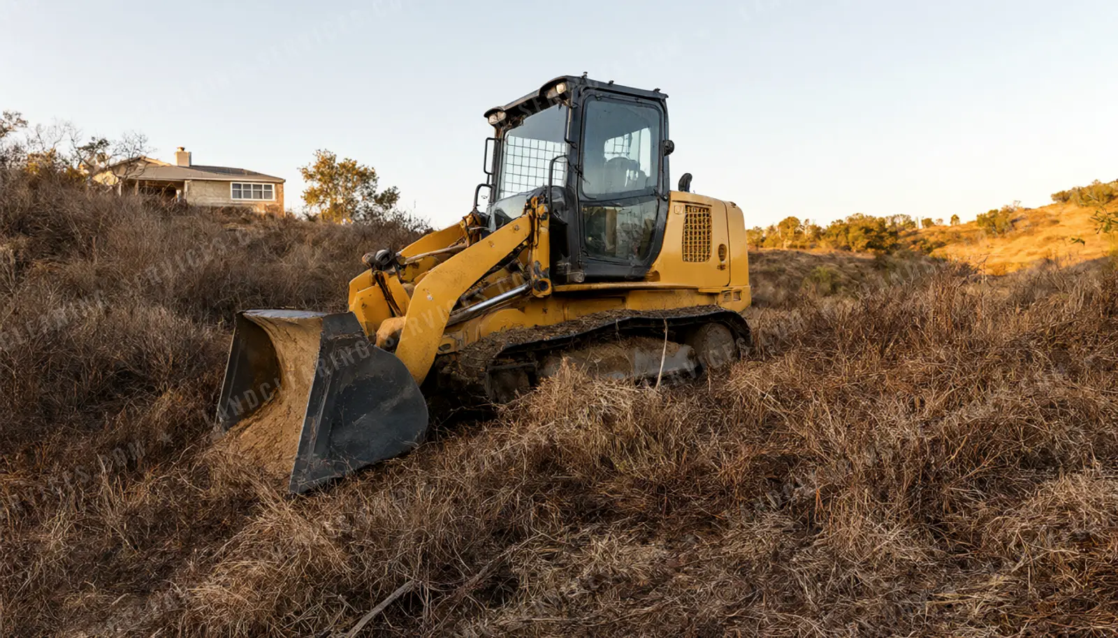 Track loader clearing dry grass and brush on a hillside below a house for defensible space, land clearing in Oregon